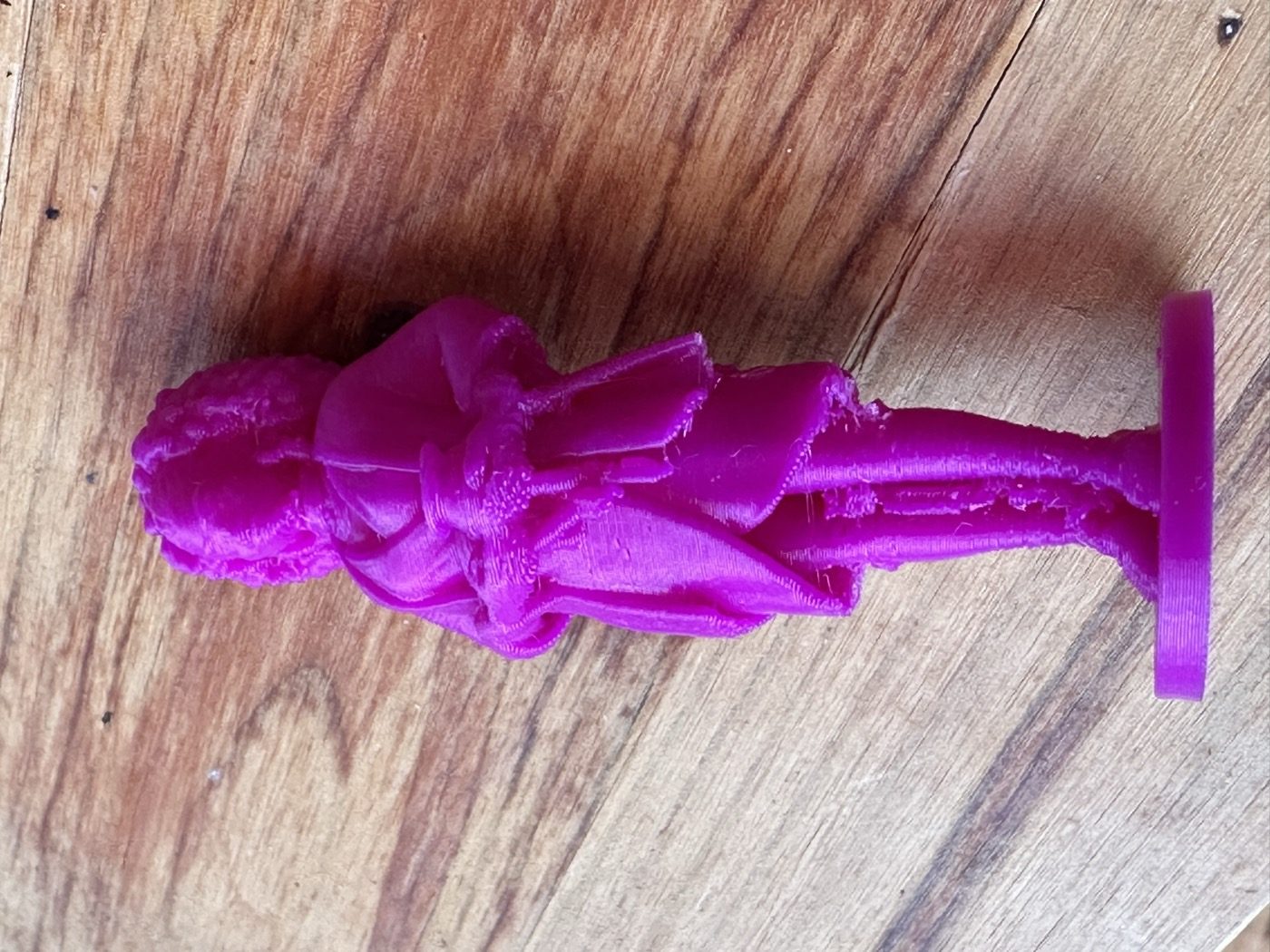 The printed 80 mm Amara figurine lying on its side on a wooden table, purple PLA, with thin support rods still attached from the dress to the hands and pot