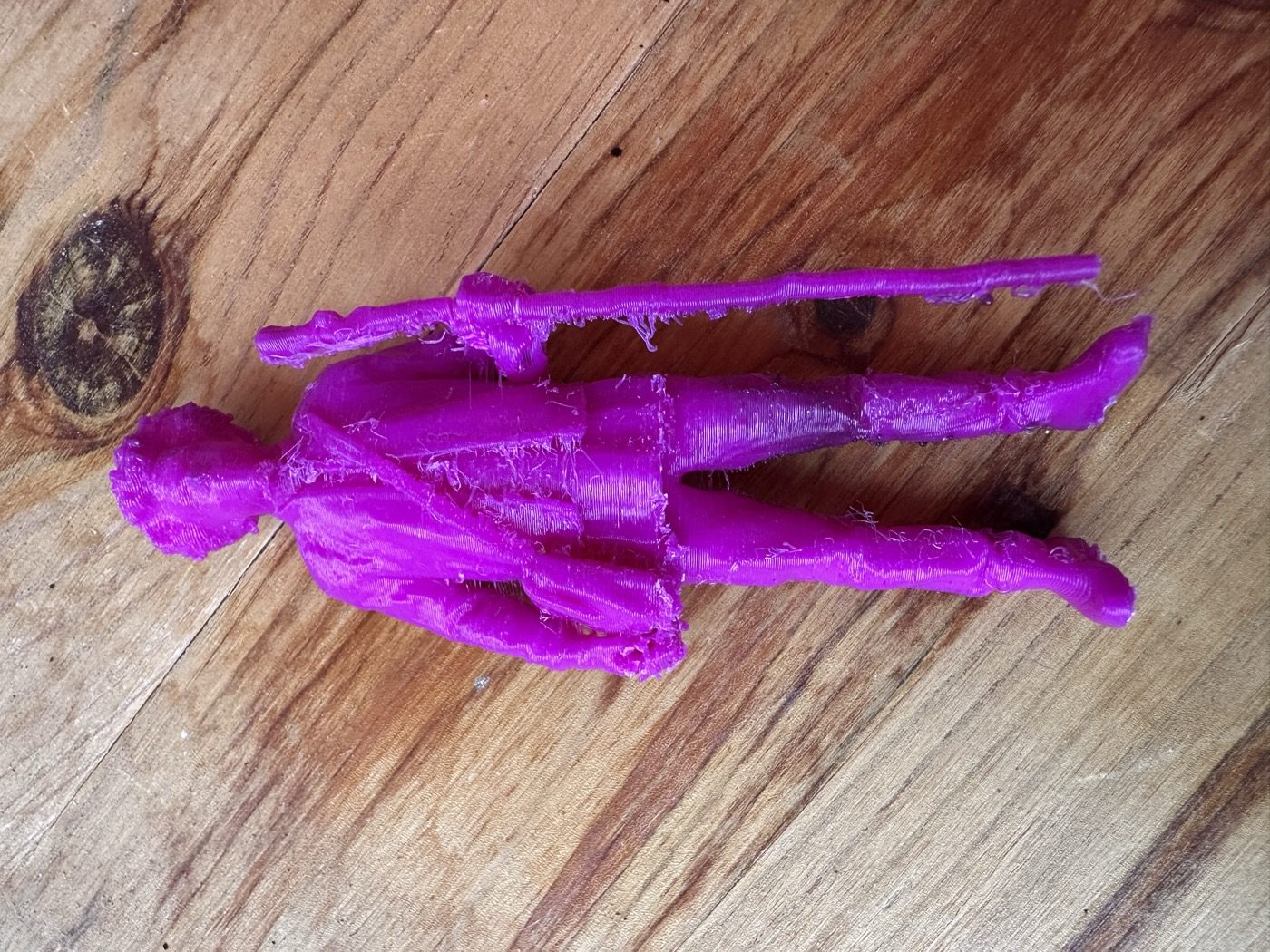 The printed 97 mm Daniel figurine lying on a wooden table: purple PLA covered in fine strings, with rough patches on the shoulder bag and along the walking staff